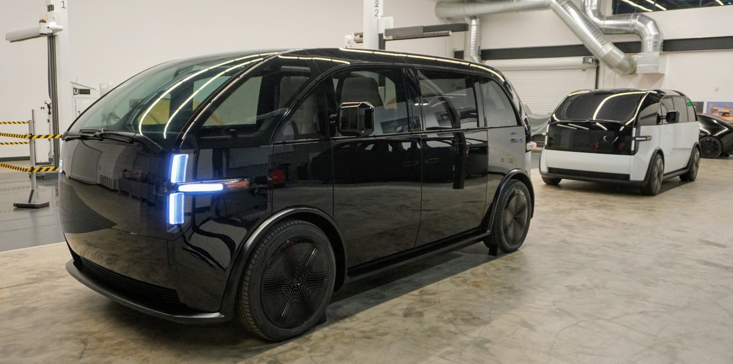 Black Canoo Lifestyle Vehicle electric van, front three-quarter view, with a white example behind