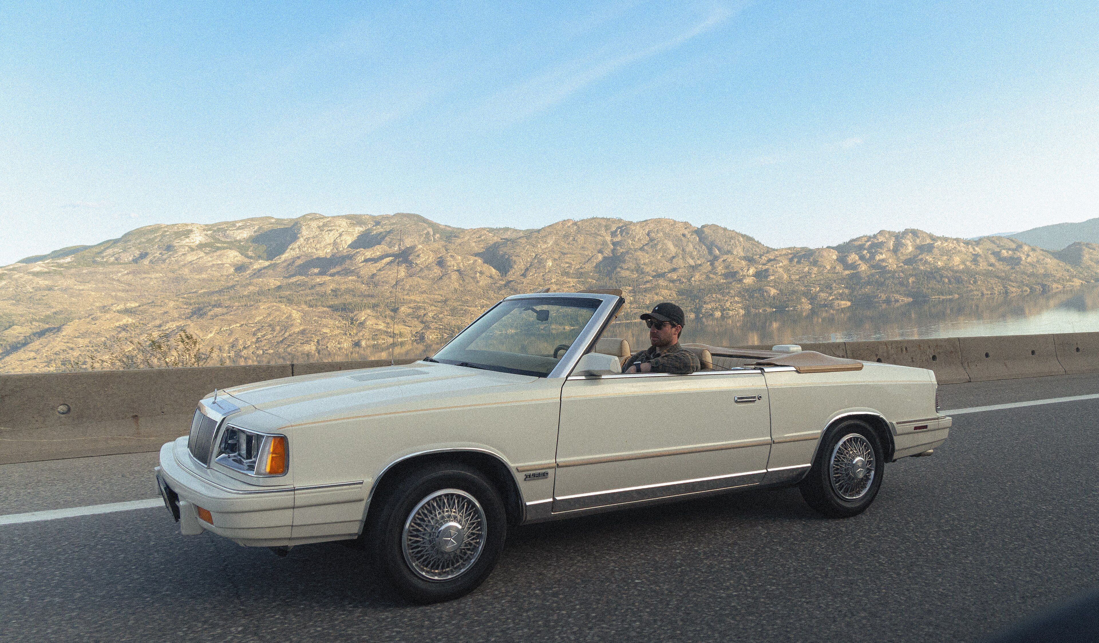 Cream 1986 Chrysler LeBaron convertible with the top down on a highway, side profile view