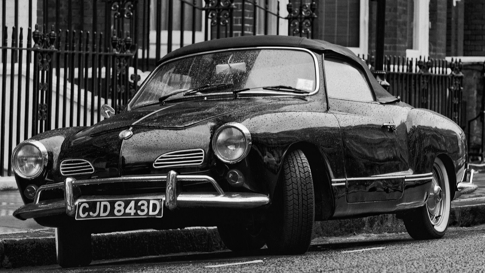 Black-and-white photograph of a 1966 Volkswagen Karmann Ghia cabriolet parked kerbside in the rain, front three-quarter view.