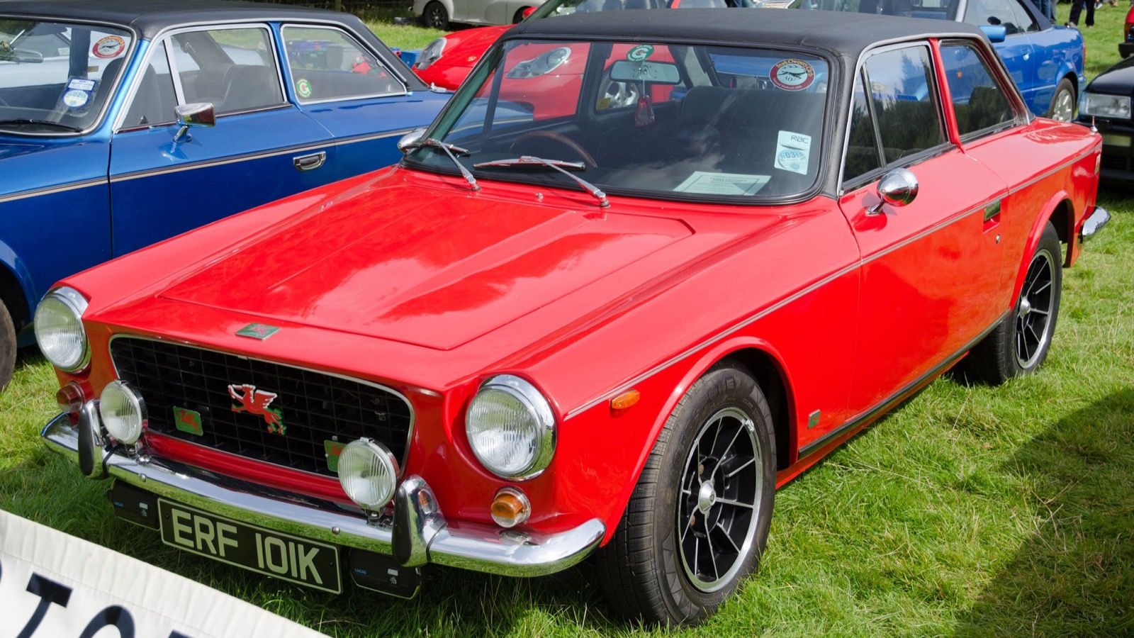 Red Gilbern Invader Mk II coupé with black vinyl roof, front three-quarter view at a classic car show.