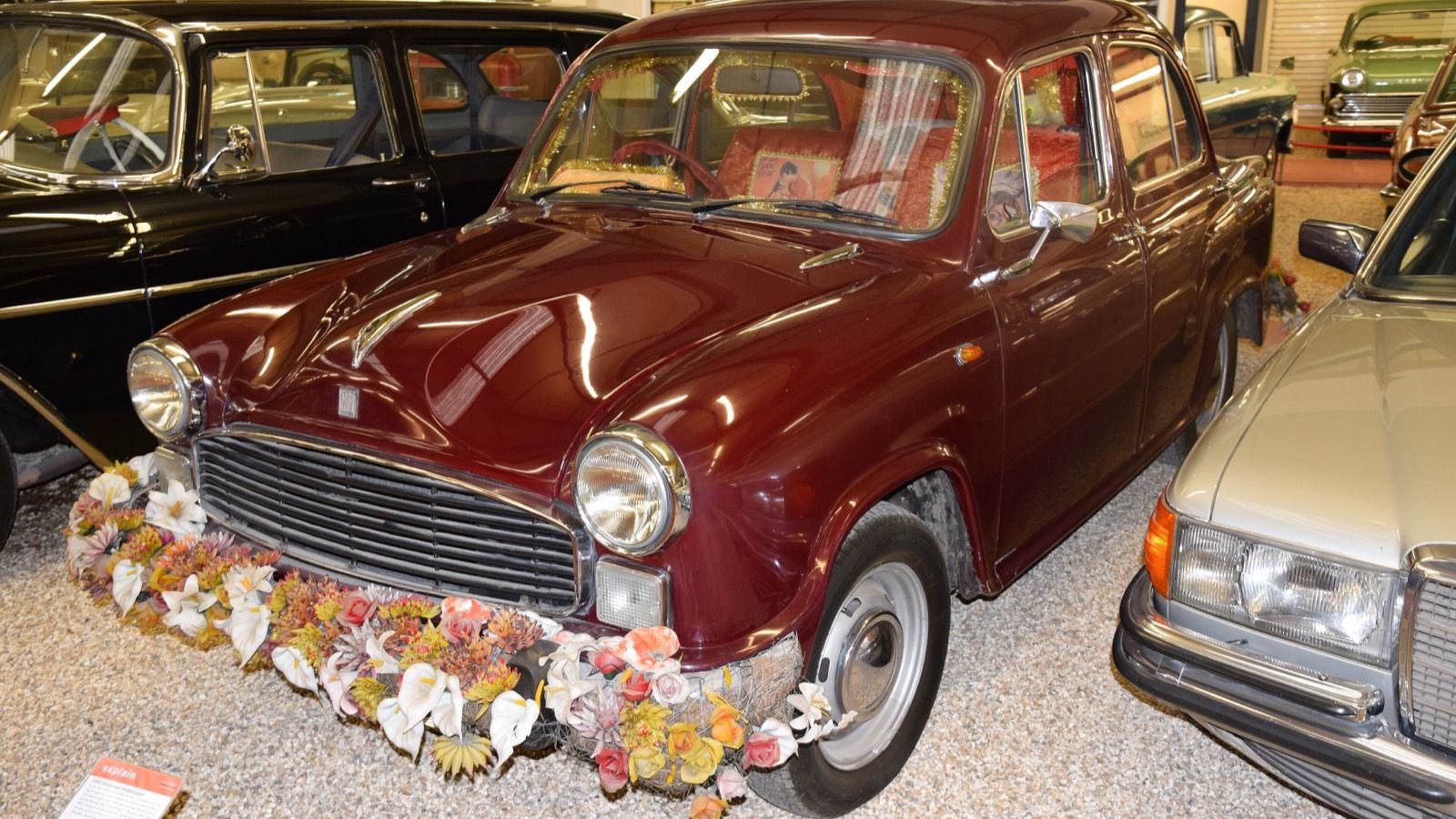 Maroon 1992 Hindustan Ambassador on museum display with floral garland across the front bumper, front three-quarter view