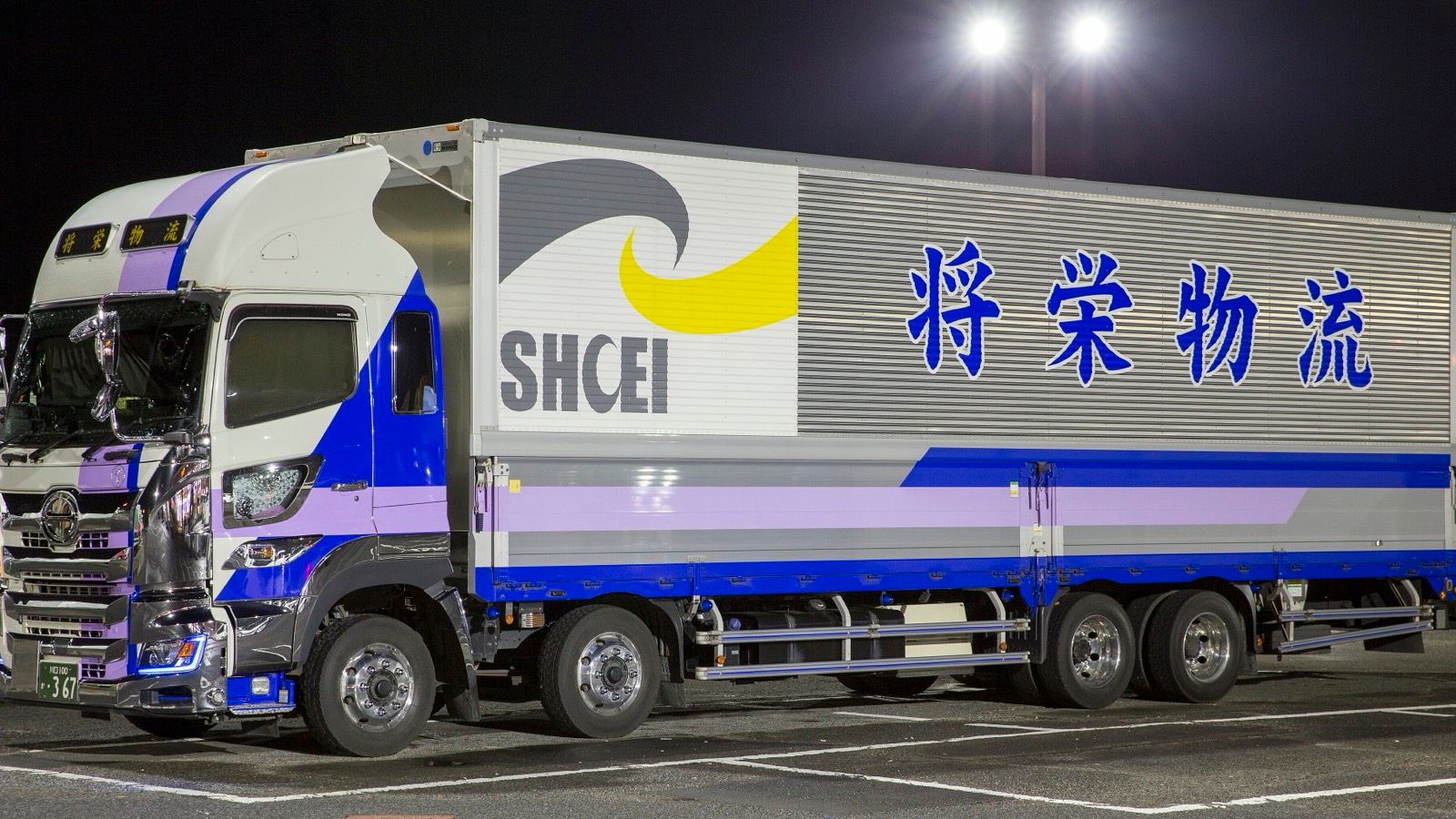 Blue and white Hino Profia wing-body truck in a lorry park at night, front three-quarter view