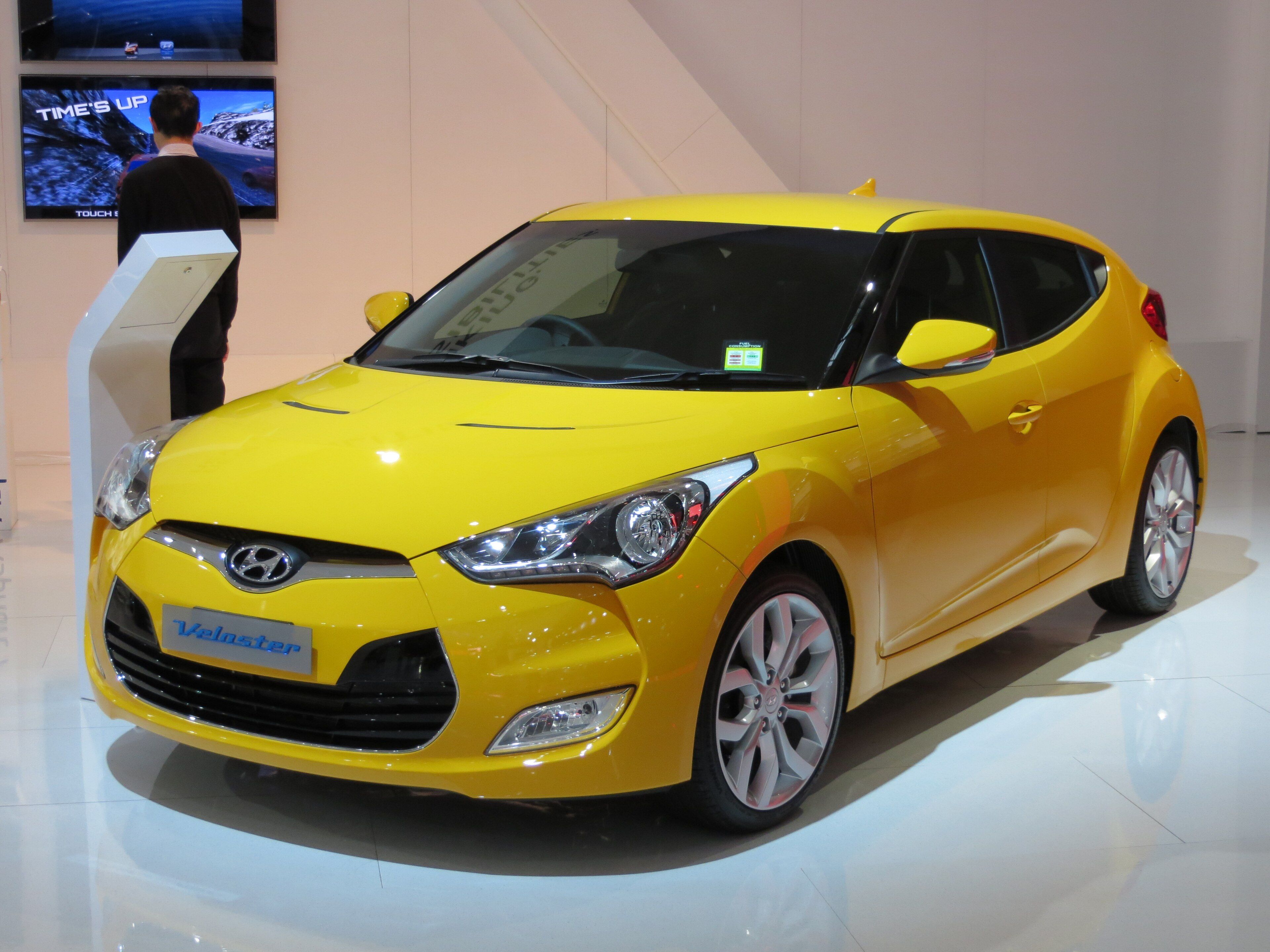 Yellow 2012 Hyundai Veloster three-door hatchback on a motor-show stand, front three-quarter view