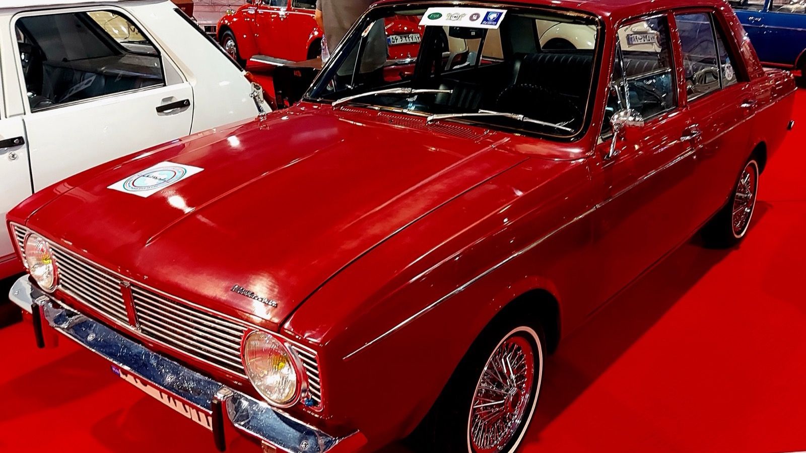 Red early Paykan saloon with the original Rootes Arrow grille on display at a classic car show, front three-quarter view