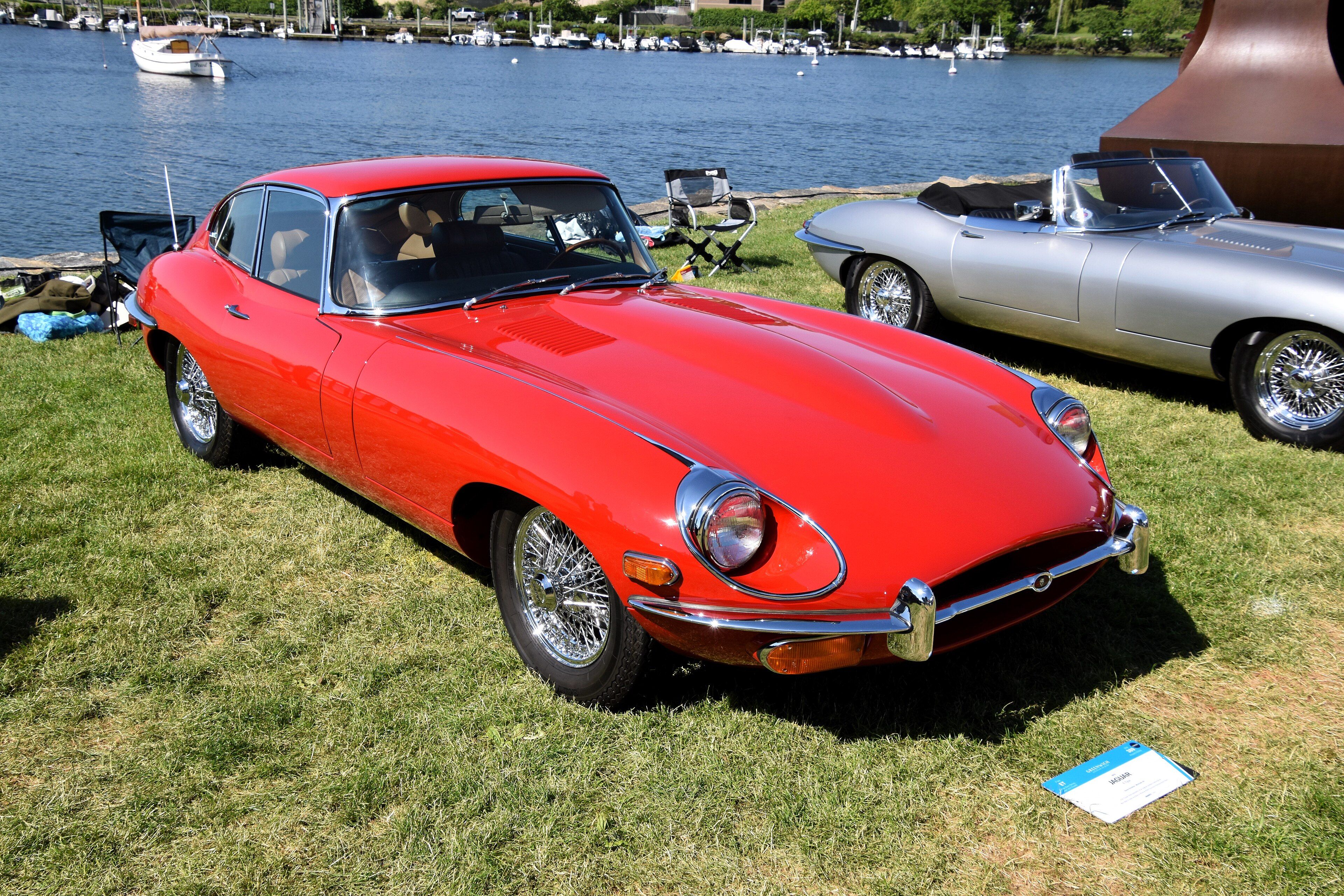 Red 1970 Jaguar E-Type 4.2 Series 2 coupe on grass at a waterside car show, front three-quarter view