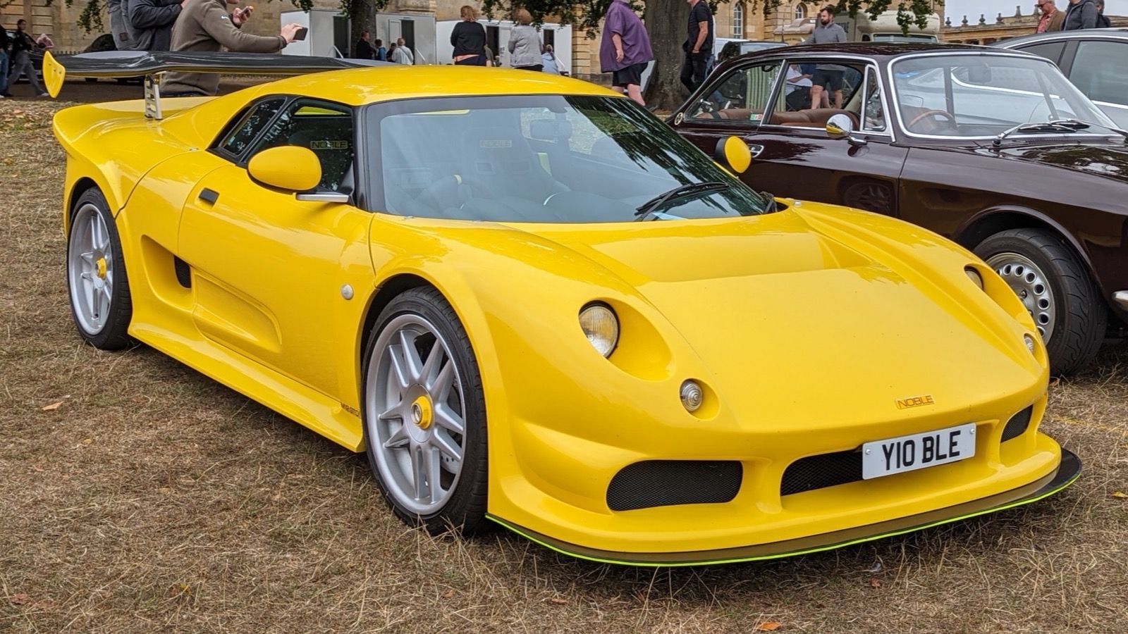 Yellow 2002 Noble M12 GTO with black rear wing parked on grass, front three-quarter view
