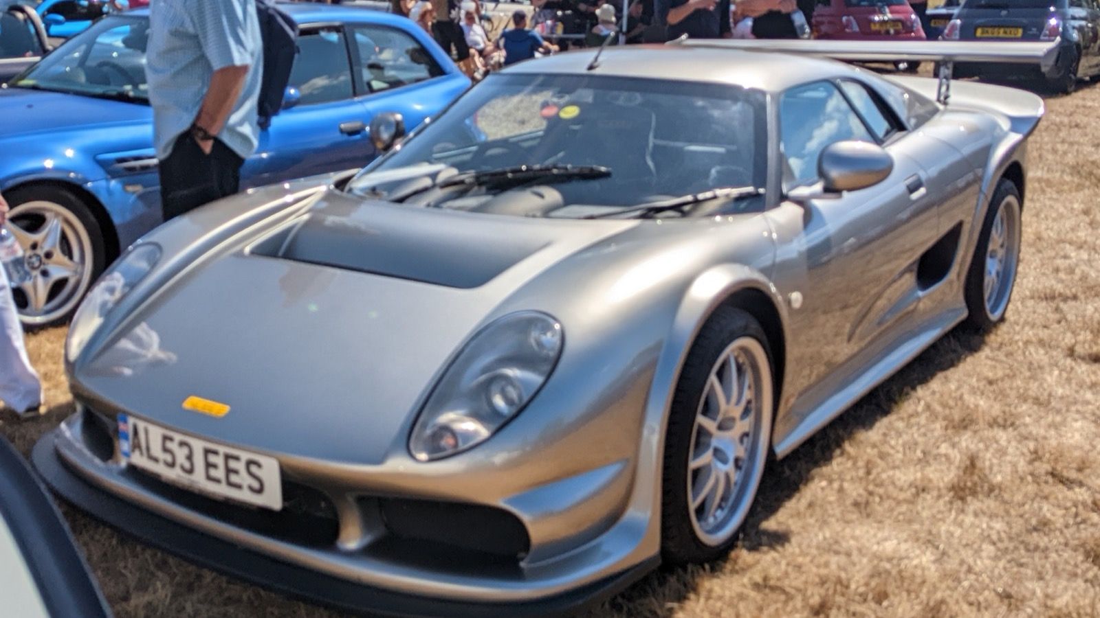 Silver 2003 Noble M12 GTO-3 parked on dry grass at a car show, front three-quarter view