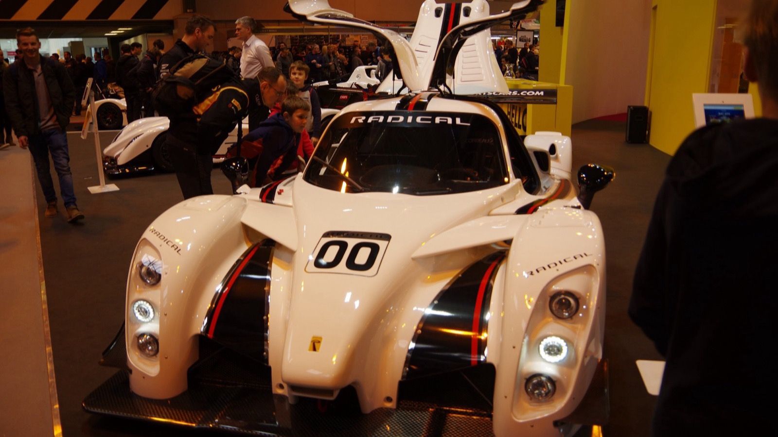 White Radical RXC Turbo 500 coupe with gullwing doors raised on an indoor show stand, front three-quarter view