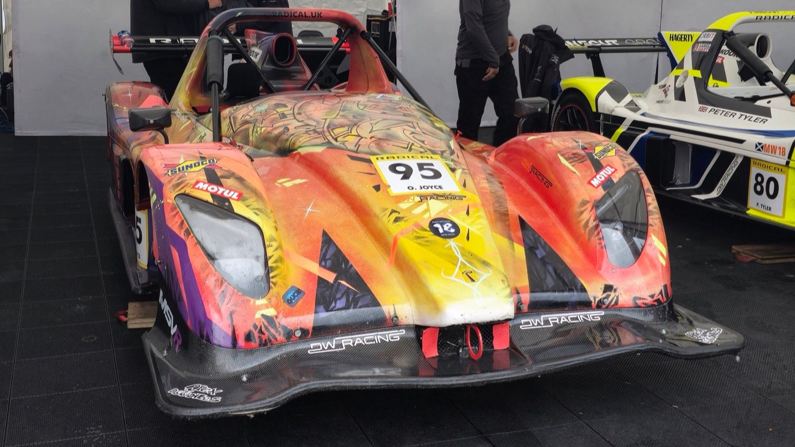 Orange and yellow liveried 2024 Radical SR3 race car in the paddock, front three-quarter view